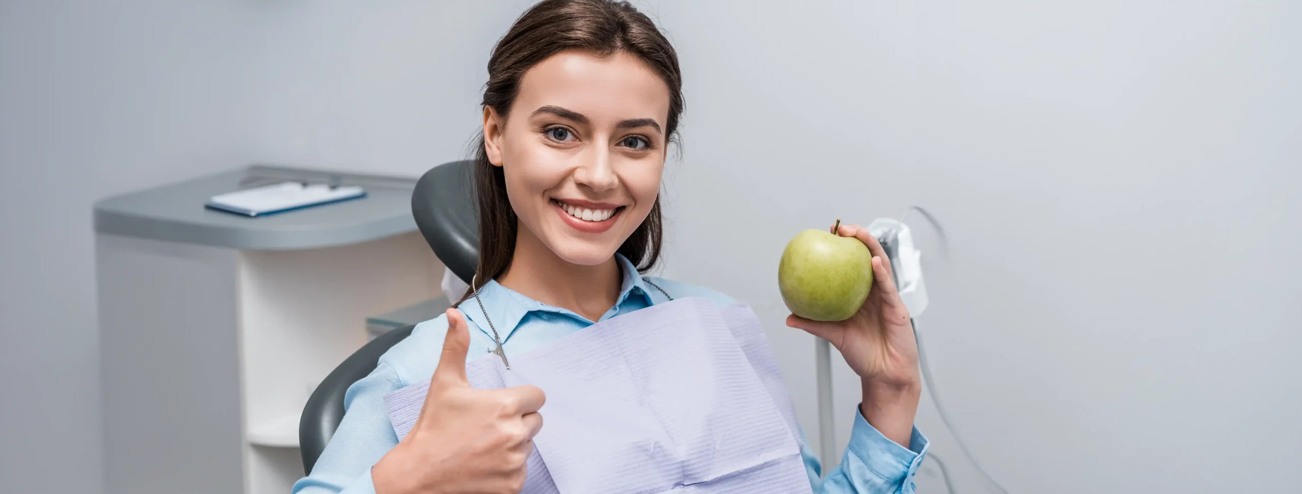 Woman sitting in a dental chair holding a green apple and giving a thumbs up