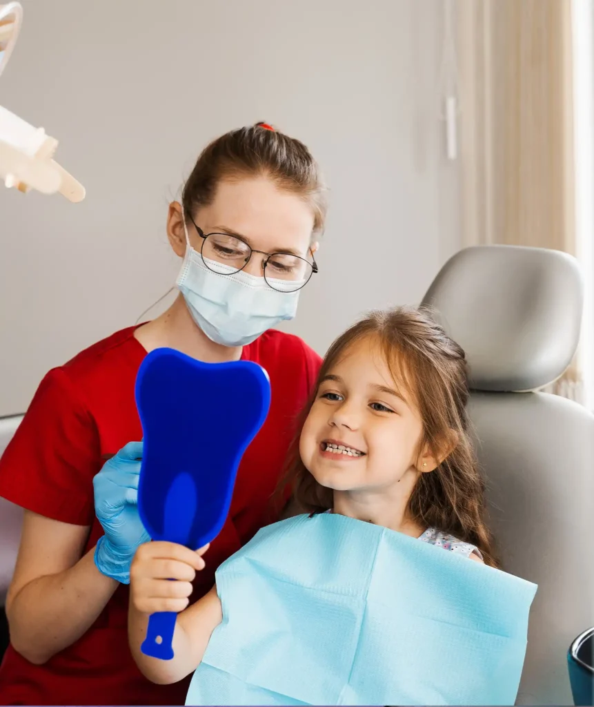 Masked dental professional holding a tooth-shaped mirror beside a smiling young girl in the dental chair