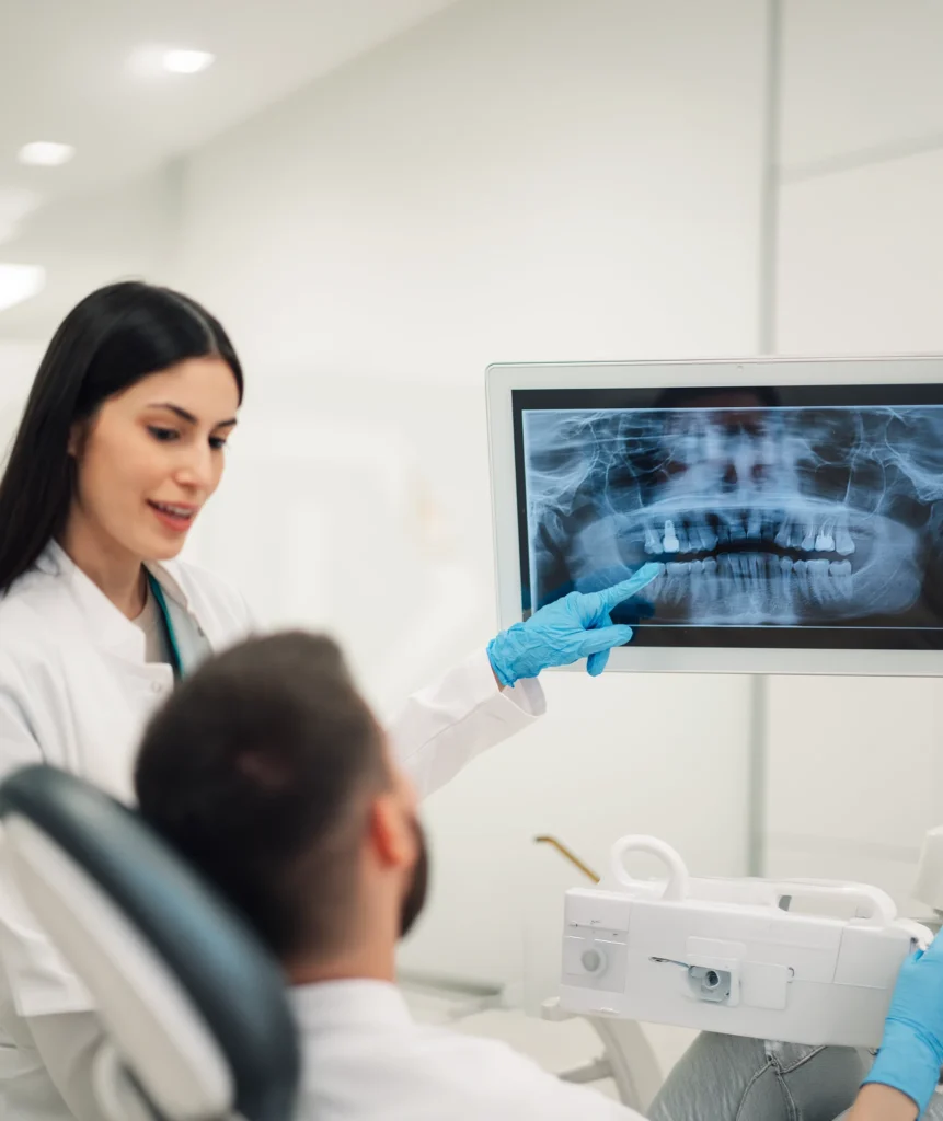 Dentist pointing at a full panoramic jaw X-ray displayed on a large screen while a patient sits chairside