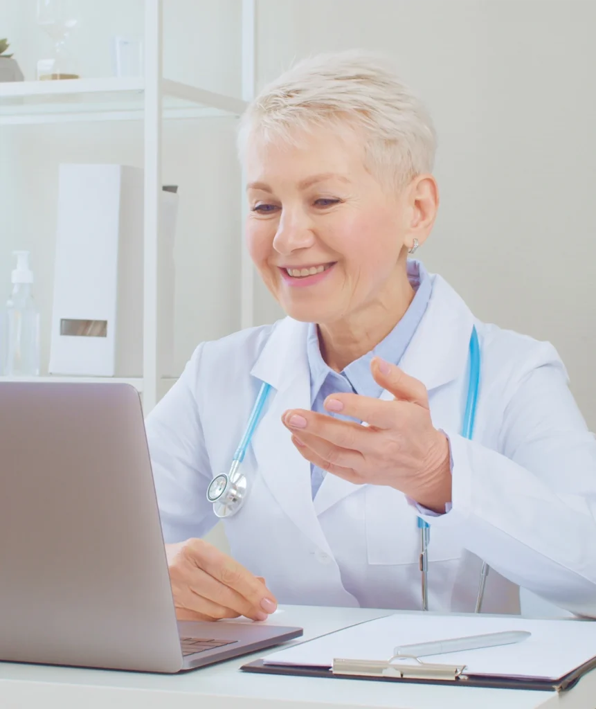 Older female doctor in a white coat smiling while gesturing during a digital consultation on a laptop