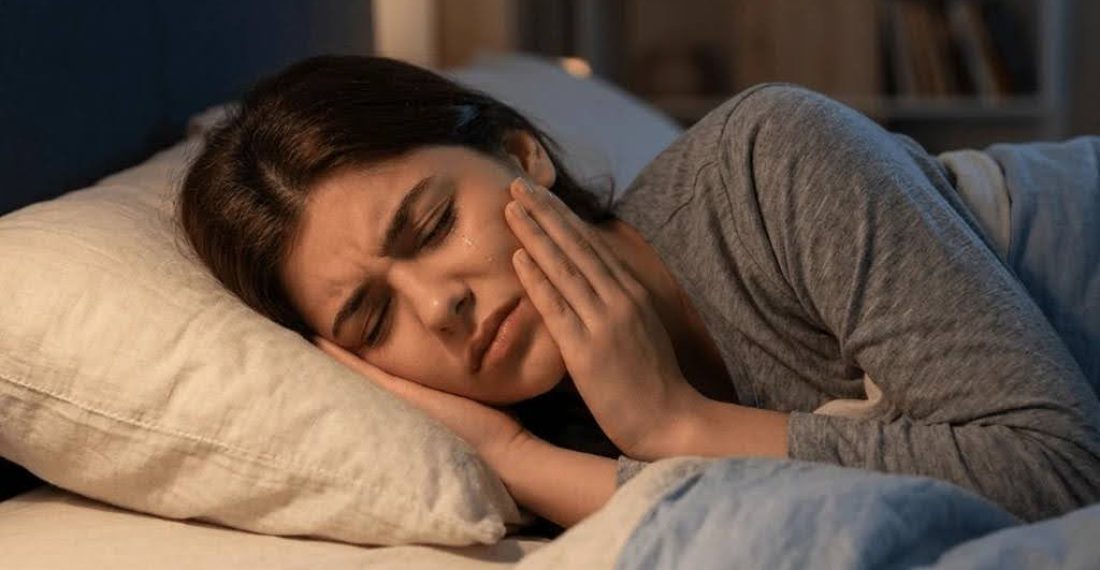 A woman lying in bed at night, looking distressed and holding her cheek in pain, indicating symptoms of a painful toothache.